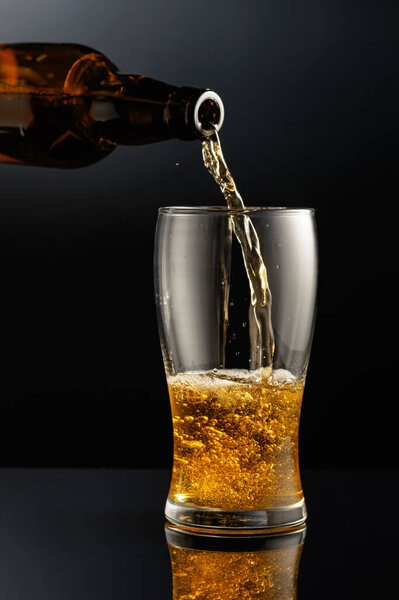 Pouring beer from a bottle into a glass. Glass of beer on a black reflective background.