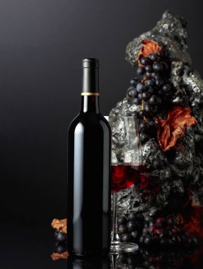 Bir şişe kırmızı şarap. Arka planda kaba gri taş ve mavi üzümler var. Boşluğu kopyala.