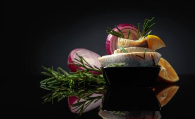 Limonlu, kırmızı soğanlı ve biberiyeli ringa fileto. Siyah yansıtıcı arka planda ringa balığı olan siyah bir tabak. Boşluğu kopyala.