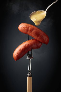 Boiled sausages with mustard on a fork. Hot sausages with smoke on a black background.