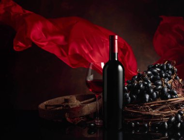 Siyah yansıtıcı arka planda üzümlü bir bardak ve bir şişe kırmızı şarap. Kırmızı saten perde rüzgarda dalgalanıyor. Boşluğu kopyala.