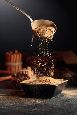 Tarçın tozu siyah bir kaseye dökülür. Arka planda mutfak aletleri ve tarçın çubukları var..