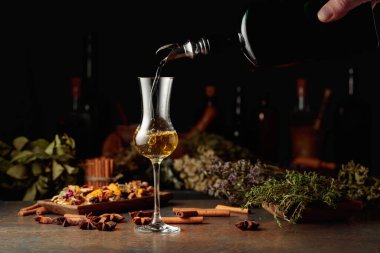 Sarı bitkisel likör ya da merhem eski bir şişeden bardağa dökülür. Kurutulmuş otlar, çiçekler, baharatlar ve eski mutfak aletleri. Bitkisel ilaç kavramı.