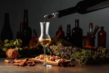 Sarı bitkisel likör ya da merhem eski bir şişeden bardağa dökülür. Kurutulmuş otlar, çiçekler, baharatlar ve eski mutfak aletleri. Bitkisel ilaç kavramı.