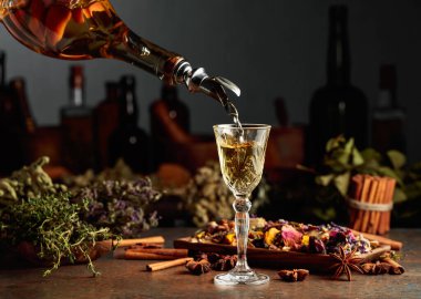 Sarı bitkisel likör ya da karışım eski bir şişeden bardağa dökülür. Kurutulmuş otlar, çiçekler, baharatlar ve eski mutfak aletleri. Bitkisel ilaç kavramı.