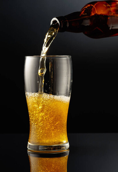 Pouring beer from a bottle into a glass. Glass of beer on a black reflective background.