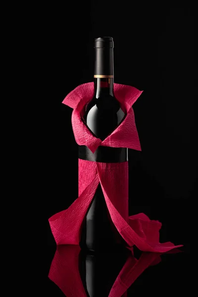 Bottle of red wine wrapped in crepe paper on a black background. The bottle looks like a woman in a red evening dress.
