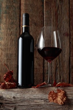 Kurumuş asma yaprakları olan eski ahşap bir masada şişe ve bir bardak kırmızı şarap..