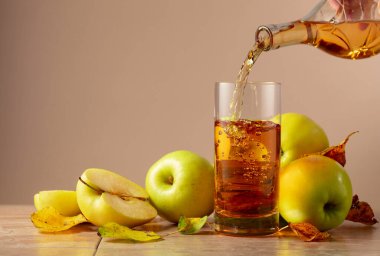 Elma suyu şişeden bardağa dökülür. Seramik bir masada olgun elmalar ve kurumuş yapraklar. Kopyalama alanı olan bej arkaplan.