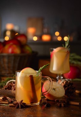 Elma dilimleri, tarçın, biberiye ve kahverengi bir kırsal masada cam bardakta anason yıldızlarıyla pişirilmiş elma şarabı. Geleneksel sıcak bayram içeceği. Karanlık bir arka planda mutfak aletleri ve yanan mumlar..