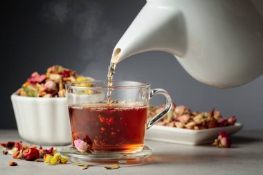 Bitki çayı bardağa dökülür. Kurumuş otlar ve çiçekler taş masaya saçılmış..