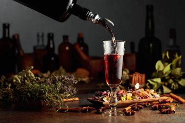 Esmer bitkisel likör ya da merhem eski bir şişeden bardağa dökülür. Kurutulmuş otlar, çiçekler, baharatlar ve eski mutfak aletleri. Bitkisel ilaç kavramı.