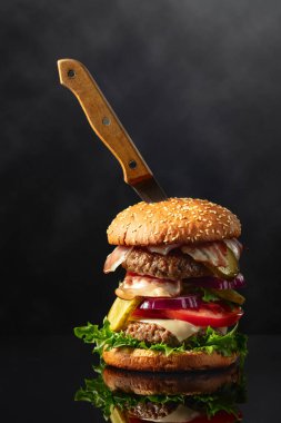 Siyah yansıtıcı arka planda bıçaklı taze lezzetli bir burger. Domatesli hamburger, soğan, korunmuş salatalık, salata, peynir, sığır eti ve pastırma..