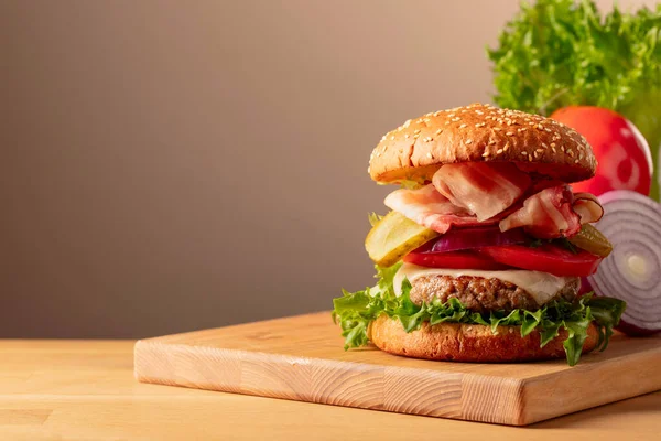 Fresh tasty burger on a wooden cutting board. Burger with tomato, onion, preserved cucumber, salad, cheese, beef cutlet, and bacon. Copy space.