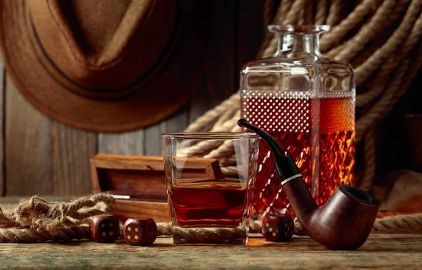 Whiskey, smoking pipe, and dice on a old wooden table. In the background is a hemp rope and a man\'s hat.