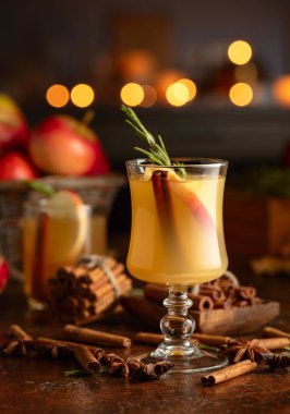 Elma dilimleri, tarçın, biberiye ve kahverengi bir kırsal masada cam bardakta anason yıldızlarıyla pişirilmiş elma şarabı. Geleneksel sıcak bayram içeceği. Karanlık bir arka planda mutfak aletleri ve yanan mumlar..