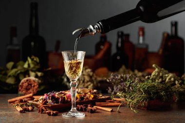 Sarı bitkisel likör ya da karışım eski bir şişeden bardağa dökülür. Kurutulmuş otlar, çiçekler, baharatlar ve eski mutfak aletleri. Bitkisel ilaç kavramı.