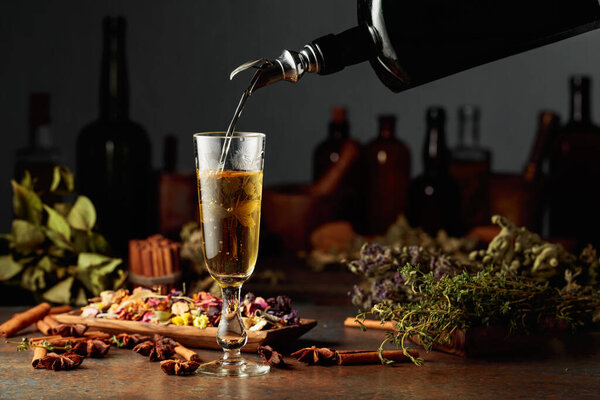 Yellow herbal liquor or mixture is poured from a vintage bottle into a glass. On a table dried herbs, flowers, spices, and old kitchen utensils. Concept of herbal medicine.