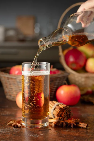Apple cider is poured from a bottle into a glass. Fresh drink with apples, cinnamon, and anise on a kitchen table.