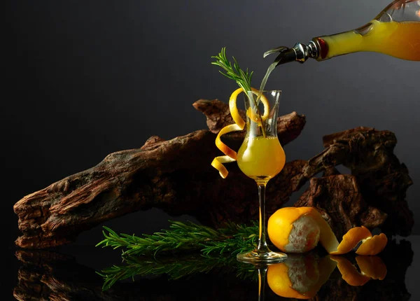 黒い反射の背景に伝統的なイタリアのリキュールリムチェッロ 古いスナッグの背景にローズマリーツイッグを施したレモン酒のグラス 液体をガラスに注入する — ストック写真