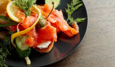 Dereotlu füme somon, salatalık, kapari, yeşil soğan ve siyah tabakta limon..