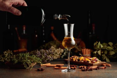 Sarı bitkisel likör ya da merhem eski bir şişeden bardağa dökülür. Kurutulmuş otlar, çiçekler, baharatlar ve eski mutfak aletleri. Bitkisel ilaç kavramı.