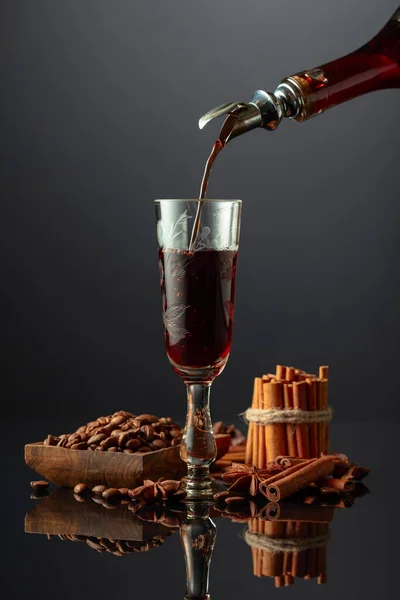Coffee liquor is poured from a bottle into a glass. Coffee beans, cinnamon sticks, and anise are scattered on the black reflective background.