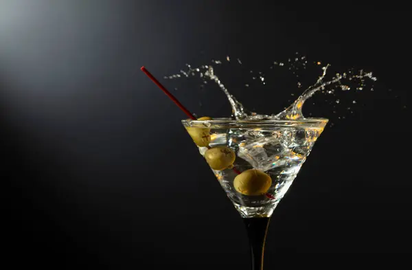 Glass of classic dry martini cocktail with green olives on a black background. Martini with splashes. Free space for your text.