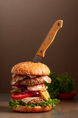 Kahverengi zemin üzerinde bıçak olan taze bir burger. Domatesli burger, soğan, korunmuş salatalık, salata, peynir, sığır pirzolası ve pastırma. Boşluğu kopyala.