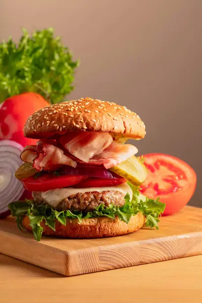 Fresh tasty burger on a wooden cutting board. Burger with tomato, onion, preserved cucumber, salad, cheese, beef cutlet, and bacon. Copy space.
