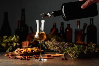 Sarı bitkisel likör ya da merhem eski bir şişeden bardağa dökülür. Kurutulmuş otlar, çiçekler, baharatlar ve eski mutfak aletleri. Bitkisel ilaç kavramı.