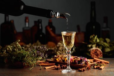 Sarı bitkisel likör ya da karışım eski bir şişeden bardağa dökülür. Kurutulmuş otlar, çiçekler, baharatlar ve eski mutfak aletleri. Bitkisel ilaç kavramı.