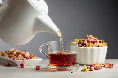 Bitki çayı bardağa dökülür. Kurumuş otlar ve çiçekler taş masaya saçılmış..