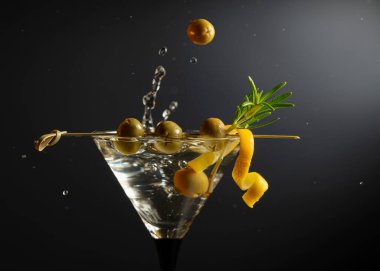 Siyah arka planda yeşil zeytinli, limon kabuklu ve biberiyeli klasik sek martini kokteyli. Sıçratmalı martini. Metnin için boş yer.