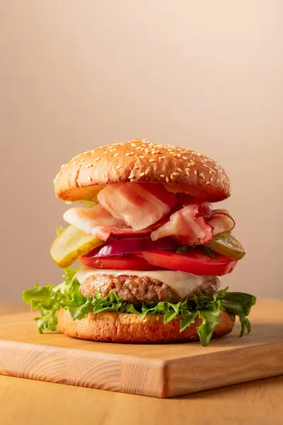 Fresh tasty burger on a beige background. Burger with tomato, onion, preserved cucumber, salad, cheese, beef cutlet, and bacon.