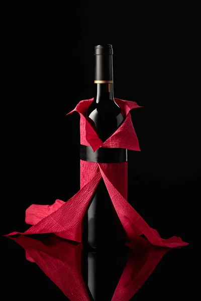 Bottle of red wine wrapped in crepe paper on a black background. The bottle looks like a woman in a red evening dress.
