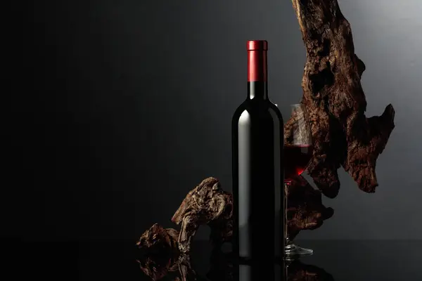 Glass and bottle of red wine. In the background old weathered snag. Black reflective background. Frontal view with space for your text.