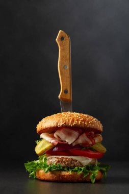 Siyah zemin üzerinde bıçak olan taze lezzetli bir burger. Domatesli hamburger, soğan, korunmuş salatalık, salata, peynir, sığır eti ve pastırma..
