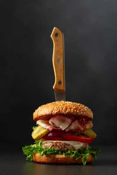 Fresh tasty burger with knife on a black background. Burger with tomato, onion, preserved cucumber, salad, cheese, beef cutlet, and bacon.