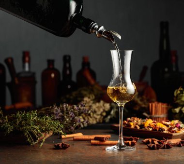 Sarı bitkisel likör ya da merhem eski bir şişeden bardağa dökülür. Kurutulmuş otlar, çiçekler, baharatlar ve eski mutfak aletleri. Bitkisel ilaç kavramı.