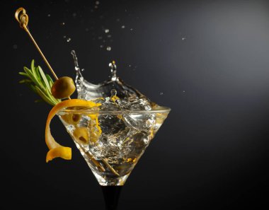 Siyah arka planda yeşil zeytinli, limon kabuklu ve biberiyeli klasik sek martini kokteyli. Sıçratmalı martini. Metnin için boş yer.