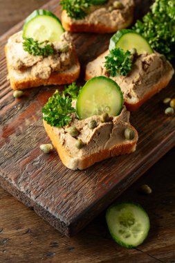 Eski bir kesme tahtasında ezmeli tost. Pate, taze salatalık, kapari ve maydanozlu sandviç..