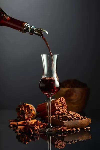 Coffee liquor is poured from a bottle into a glass. Coffee beans, cinnamon sticks, and anise are scattered on the black reflective background.