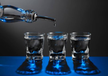 Votka şişeden koyu bir arka planda bir bardağa dökülüyor. Mavi arka ışık. Boşluğu kopyala.