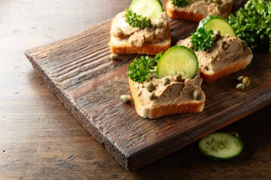 Eski bir kesme tahtasında ezmeli tost. Pate, taze salatalık, kapari ve maydanozlu sandviç..