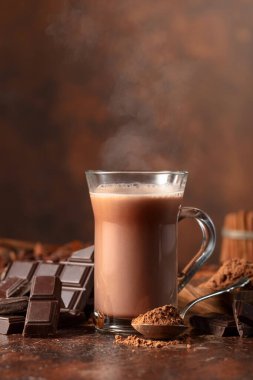 Bir parça acı çikolata, kakao tozlu kaşık ve kahverengi bir masada bir bardak kakao.. 