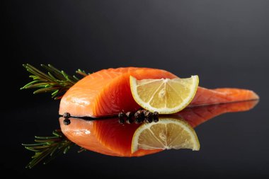 Smocked salmon piece with rosemary, lemon, and peppercorn on a black reflective background.