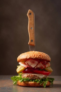 Kahverengi zemin üzerinde bıçak olan taze bir burger. Domatesli hamburger, soğan, korunmuş salatalık, salata, peynir, sığır eti ve pastırma..