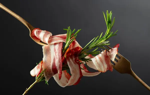 黒い背景にローズマリーが付いた乾燥した豚肉ベーコン フォークにスライスベーコン — ストック写真