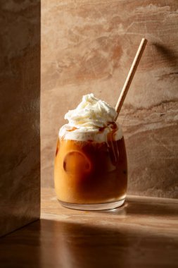 Krem şantili ve karamel soslu buzlu karamel latte, bej seramik masada ferahlatıcı ve tatlı bir kahve..